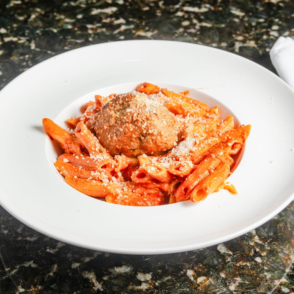 Penne pasta in creamy red sauce topped with a breaded meatball and grated cheese on a white plate.