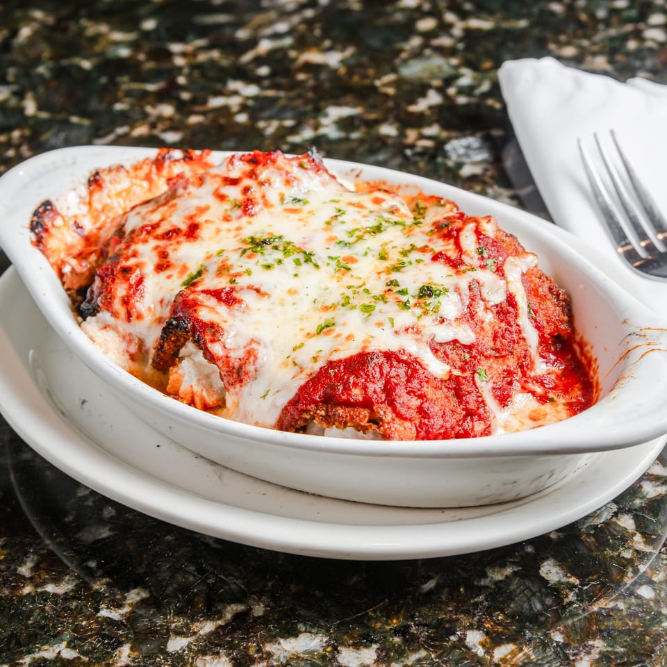 Baked eggplant Parmesan topped with melted mozzarella, tomato sauce, and herbs in a white serving dish.