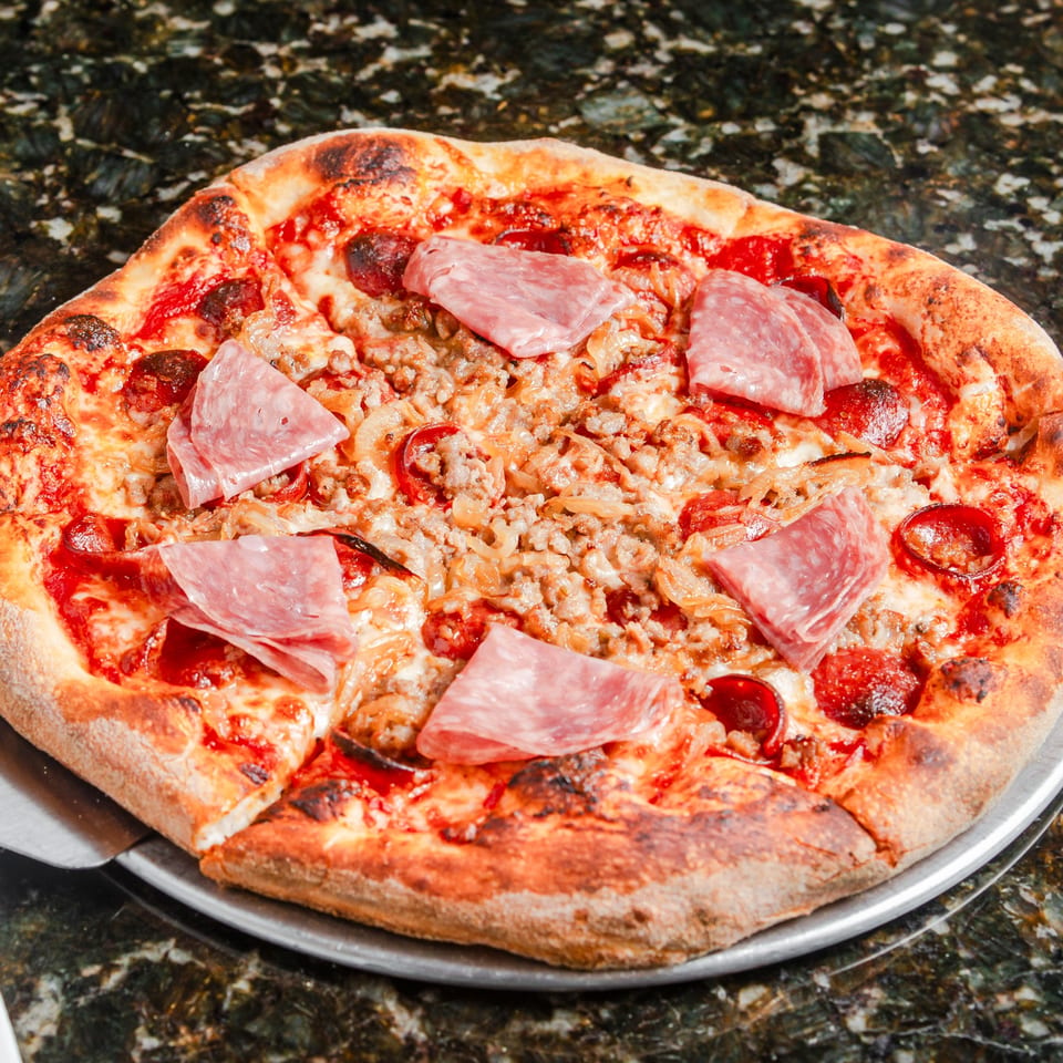 Meat-lovers pizza topped with pepperoni, sausage, onions, and folded slices of ham on a metal tray.
